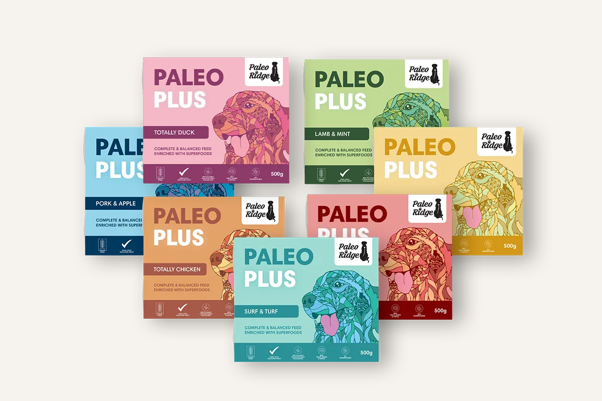 A collection of seven colorful pet food packages from Paleo Ridge, each featuring different flavors such as Totally Duck, Pork & Apple, Lamb & Mint, Totally Chicken, and Surf & Turf, all labeled "Complete & Balanced Feed Enriched with Superfoods," with stylized images of dogs and the brand logo on them. Each package is marked with a 500g weight label.